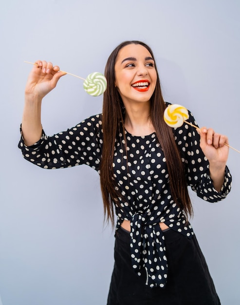 Young beauty in dotted shirt with two lollipops in hands Cheerful lady with candies over grey background