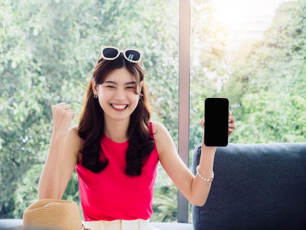 Young beauty happy smiling Asian woman portrait showing empty black blank smartphone screen and raised her fist with success gesture sit on sofa in living room with green nature summer background