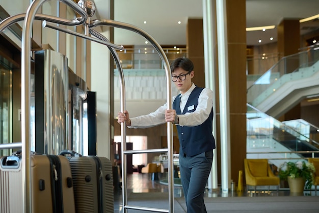 Young bellboy in uniform pushing cart with suitcases of hotel clients or guests