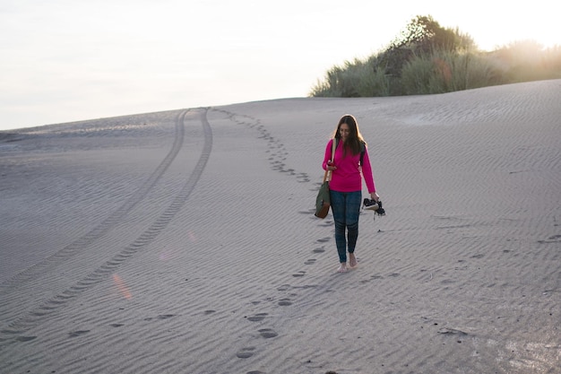 Young bespectacled Latina woman walking face forward using her cell phone on the dunes