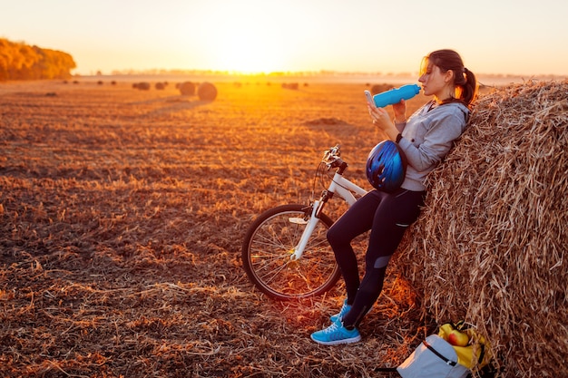 Young bicyclist having rest after a ride in autumn field at sunset. Woman drinks water by haystack and using phone to search the way