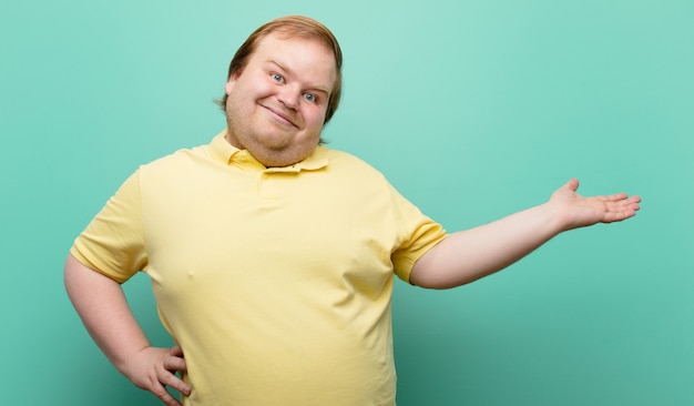 Young big size man smiling, feeling confident, successful and happy, showing concept or idea on copy space on the side against blue wall