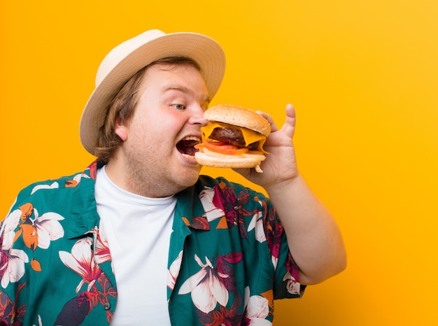 Young big size man with a cheese burger against flat wall