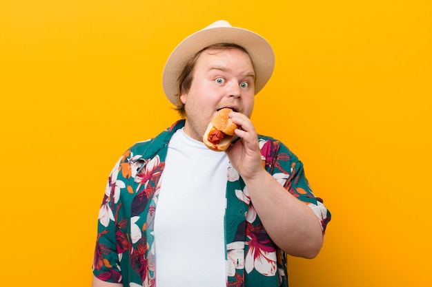 Young big size man with a hot dog against flat wall