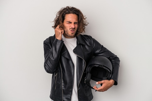 Young biker caucasian man holding a motorbike helmet isolated on gray background covering ears with hands.