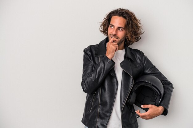 Young biker caucasian man holding a motorbike helmet isolated on gray background relaxed thinking about something looking at a copy space.