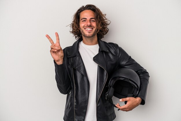 Photo young biker caucasian man holding a motorbike helmet isolated on gray background showing number two with fingers.