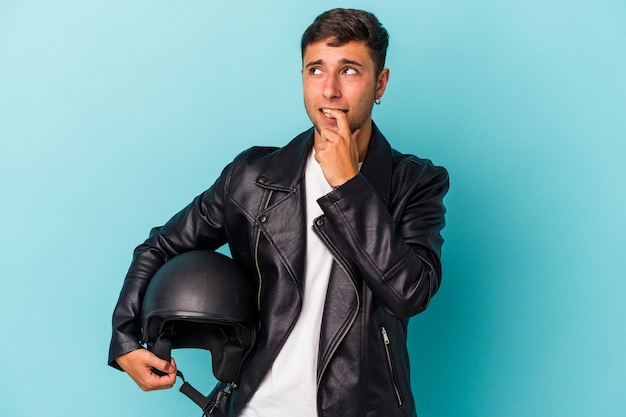 Young biker man holding helmet isolated on blue background  relaxed thinking about something looking at a copy space.