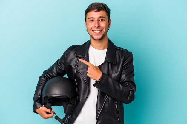 青い背景に分離されたヘルメットを保持している若いバイカーの男は、笑顔で脇を指して、空白のスペースで何かを示しています。