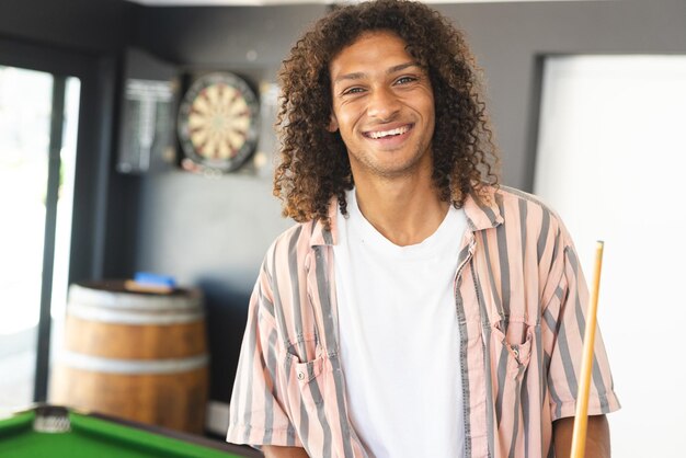 Photo young biracial man with curly hair smiles holding a pool cue