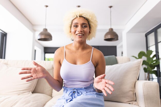 Photo young biracial woman with curly blonde hair sits on couch gesturing while talking on a video call