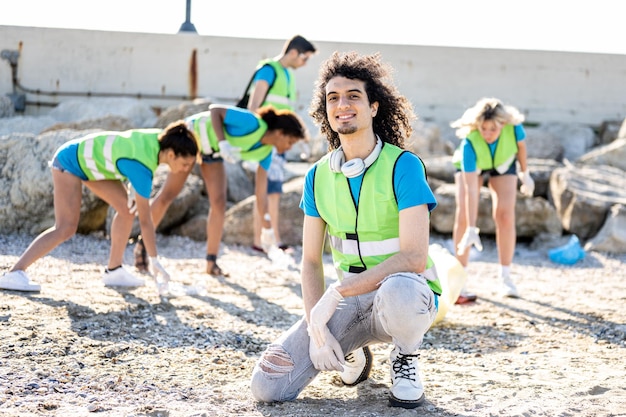 Giovane giovane birazziale che sorride alla telecamera mentre il suo lavoro di squadra pulisce la spazzatura su una spiaggia