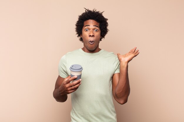 Young black afro man looking surprised and shocked, with jaw dropped holding an object with an open hand on the side