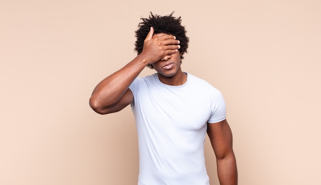 young black afro man smiling and feeling happy, covering eyes with both hands and waiting for unbelievable surprise