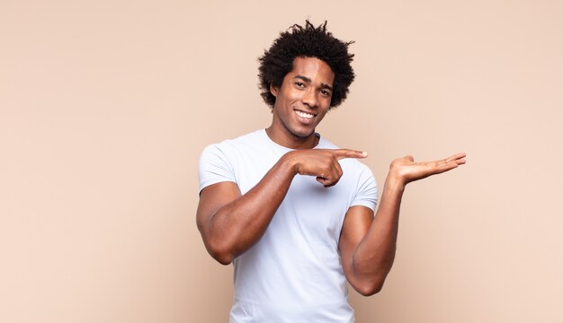 Young black afro man smiling, feeling happy, positive and satisfied, holding or showing object or concept on copy space