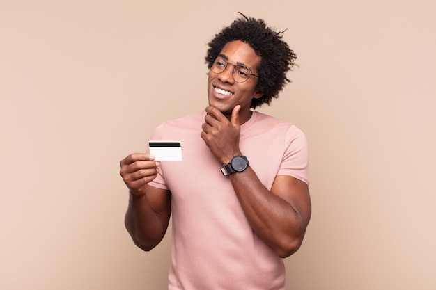 Young black afro man smiling with a happy, confident expression with hand on chin, wondering and looking to the side