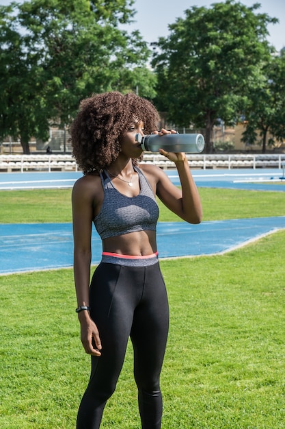 アフロヘアー黒のスポーツウェアと灰色のトップスタンディング飲料水と草の青いランニングトラックと自然光の背景の木と若い黒人アスリートの女の子