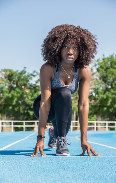 Молодая темнокожая девушка-спортсменка с афро-волосами, присевшая на корточки, готова начать гонку в черной спортивной одежде и сером топе над синей беговой дорожкой, зелеными деревьями и голубым небом на заднем плане