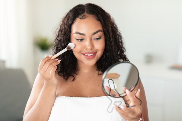 Young black body positive lady holding mirror and applying blush on face with makeup brush doing daily makeup