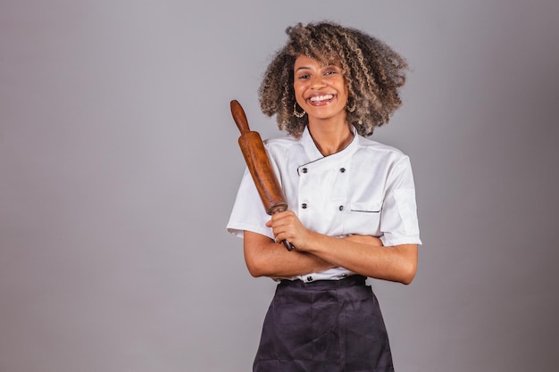 Young black Brazilian woman cook masterchef wearing restaurant uniform holding wooden rolling pin for preparing pasta bread and pizza