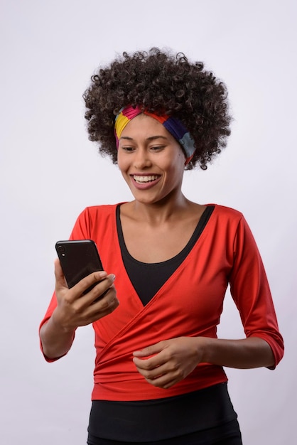 Фото Молодая чернокожая бразильская женщина, держащая и смотрящая на мобильный телефон, улыбается, нося красную блузку