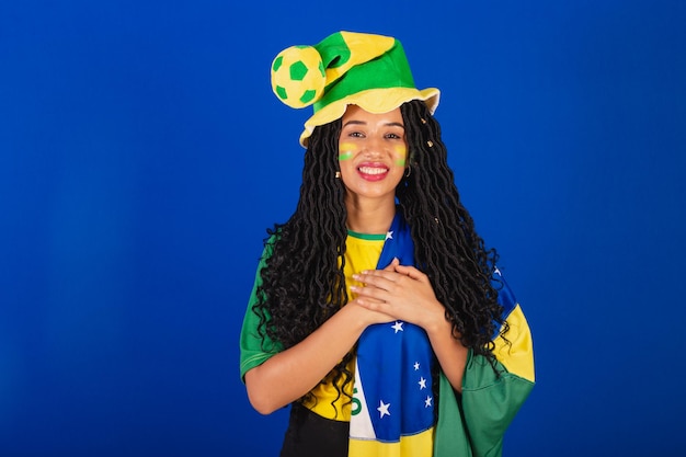 Young black Brazilian woman soccer fan with brazil flag with hand on chest gratitude