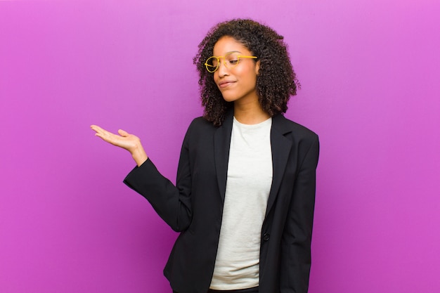 Young black business woman feeling happy and smiling casually, looking to an object or concept held on the hand on the side