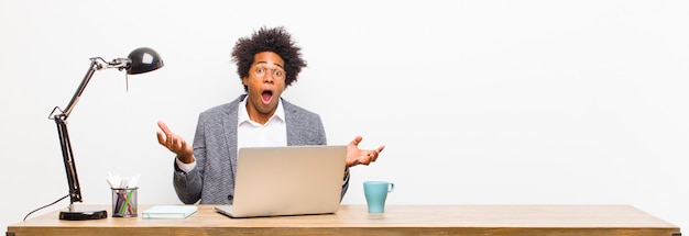 Young black businessman feeling extremely shocked and surprised, anxious and panicking, with a stressed and horrified look on a desk