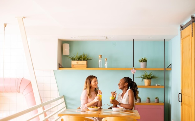 Young black and caucasian woman having good time drinking fresh juices and having healthy breakfast in the cafe