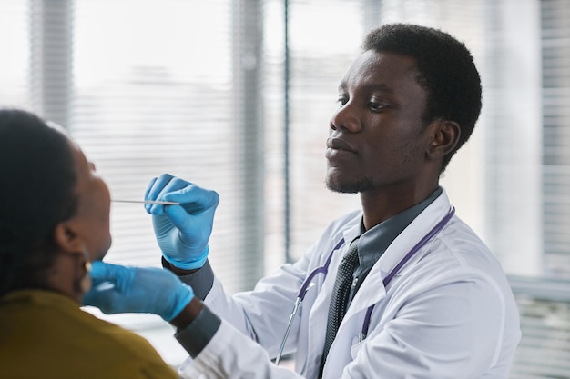Молодой черный врач консультирует пациента в клинике, проводя обследование горла на грипп