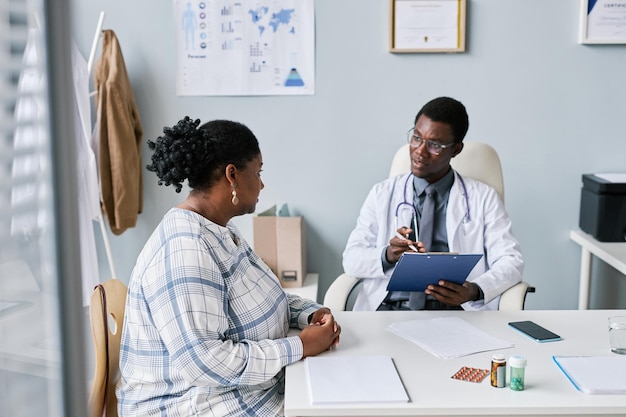 Young black doctor consulting young woman in clinic listening to symptoms