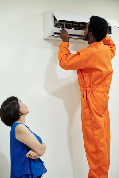 Young black engineer in orange suit providing maintenance of air conditioner while Asian female flat owner controlling his work