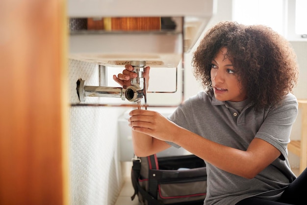 Young black female plumber sitting on the floor fixing a bathroom sink seen from doorway