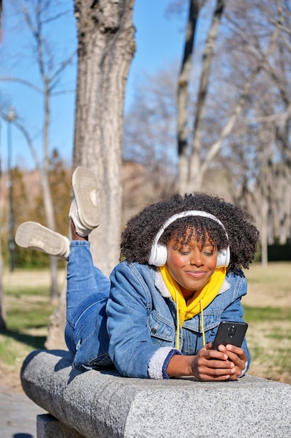 Young black girl lying on a bench listening to music wearing white headphones with her smart phone