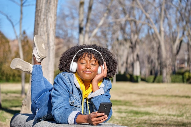 Photo young black girl lying on a bench listening to music with her eyes closed with her smart phone