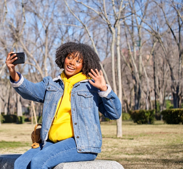 公園のベンチに座って微笑んでいる若い黒人の女の子がスマートフォンで自撮りをしている