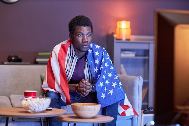 Молодой темнокожий мужчина в роли спортивного болельщика смотрит матч по телевизору дома и носит флаг США