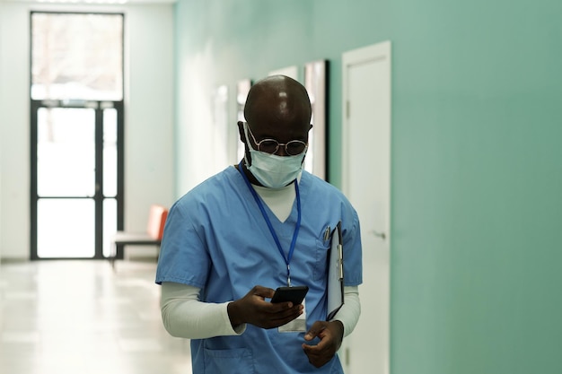 Young black man in blue uniform and protective masks texting in smartphone