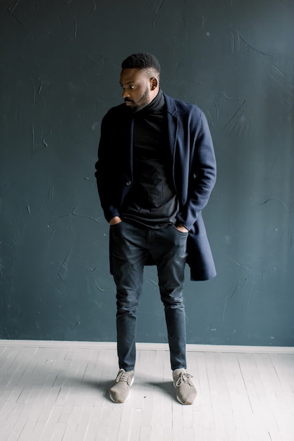 暗い背景にコートを着た若い黒人男性。プロの写真スタジオ。スタジオで黒いシャツを着た若い男のファッションの肖像画。