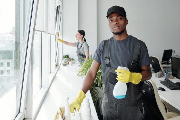 Young black man in coveralls tshirt cap and rubber gloves using detergent