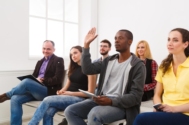 Young black man from multiethnic audience raising hand to ask question on conference, copy space