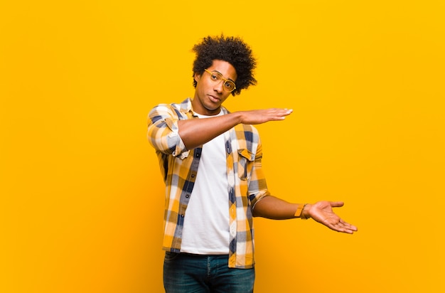 Young black man holding an object with both hands on side copy space, showing, offering or advertising an object against orange wall