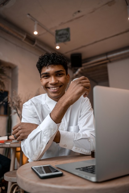 A young black man is sitting at the table with a laptop, drinking tea and using a smartphone. The guy in the cafe. Work outside the office, remote work or study concept. Vertical photo.