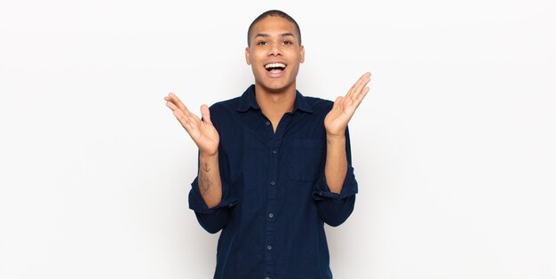 Young black man looking happy and excited, shocked with an unexpected surprise with both hands open next to face