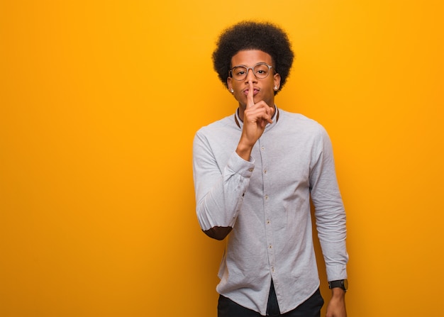 Young black man over an orange wall keeping a secret or asking for silence