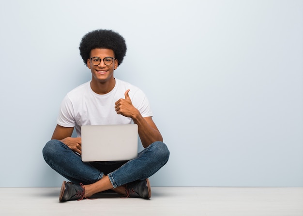 Giovane uomo di colore che si siede sul pavimento con un computer portatile che sorride e che alza pollice in su