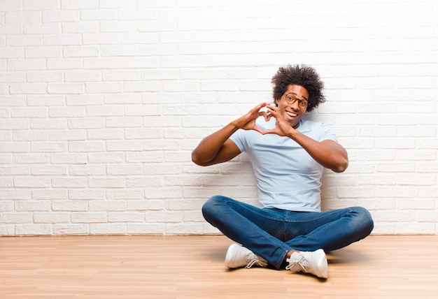 Giovane uomo di colore che sorride e sentirsi felice, carino, romantico e innamorato, facendo a forma di cuore con entrambe le mani seduto sul pavimento a casa