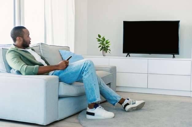 Young black man watching tv with empty screen resting on sofa in living room interior mockup