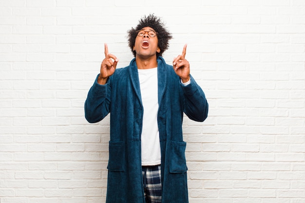 Photo young black man wearing pajamas with gown feeling awed and open mouthed pointing upwards with a shocked and surprised look against brick