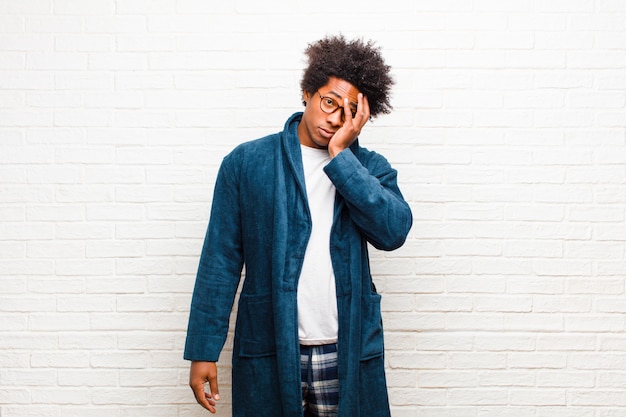 Young black man wearing pajamas with gown feeling bored, frustrated and sleepy after a tiresome, dull and tedious task, holding face with hand   brick wall